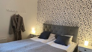 A neatly made bed with a grey headboard and white bedding, two bedside lamps, a robe hanging on a hook, and a floral-patterned wall. - 5 Star Holiday Home in Uddevalla (Uddevalla)