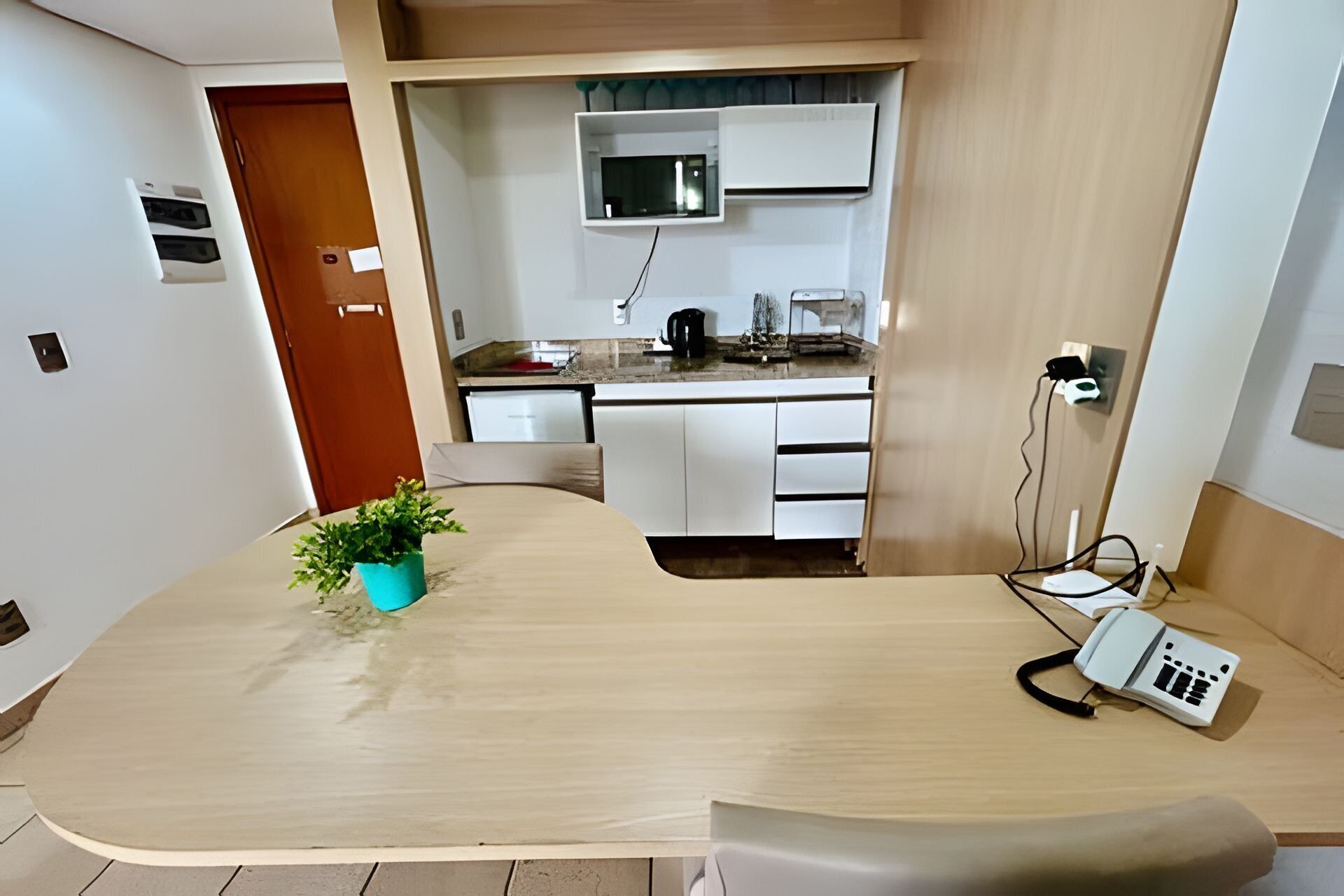 Una cocina moderna con armarios blancos, un microondas y una cafetera. Hay un escritorio de madera con una planta y un cargador de teléfono.