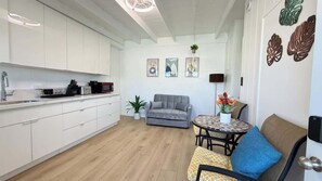 A modern kitchen with white cabinets, a grey sofa, a round table with a plant, and artwork on the wall. - Rycroft Residence 1BR Apt with AC & Full Kitchen (Honolulu)