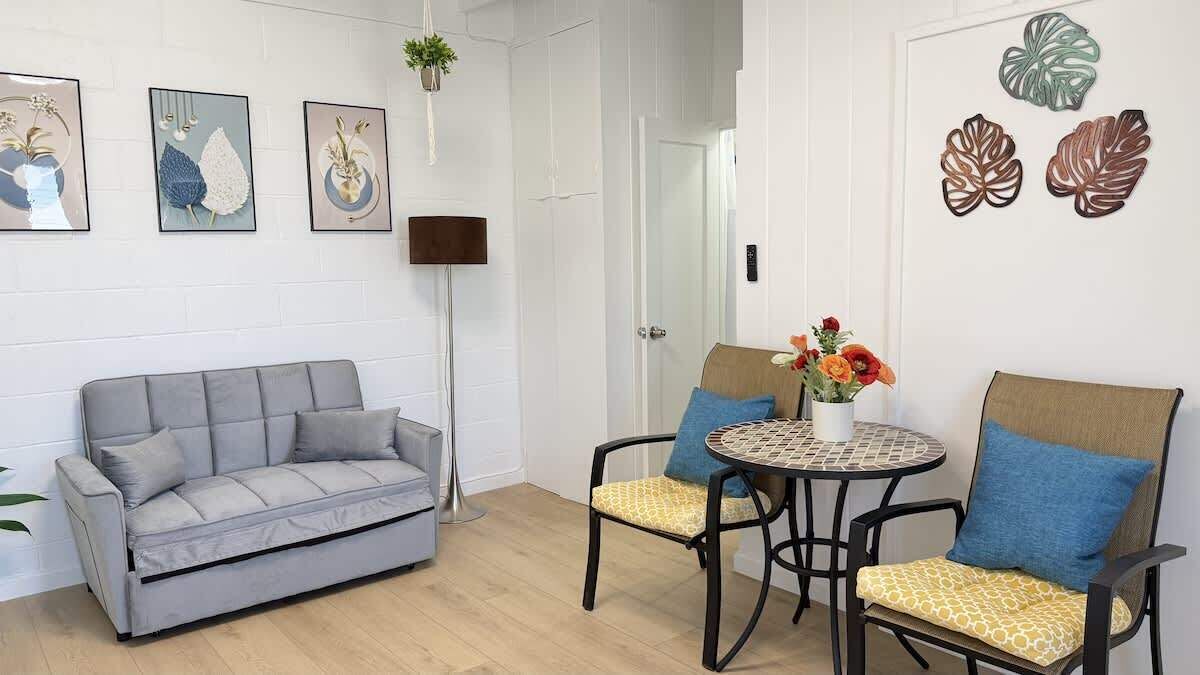 A living room with a grey sofa, two chairs with cushions, a small round table with a vase of flowers, and wall art.