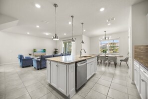 A modern kitchen with white cabinetry, a central island, pendant lighting, and a dining area with a view. - 5 BD/ 4 BA/ Solterra (7247OL) (Davenport)