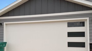 Un portail de garage de couleur blanche, une poubelle verte et une maison avec un revêtement extérieur gris.