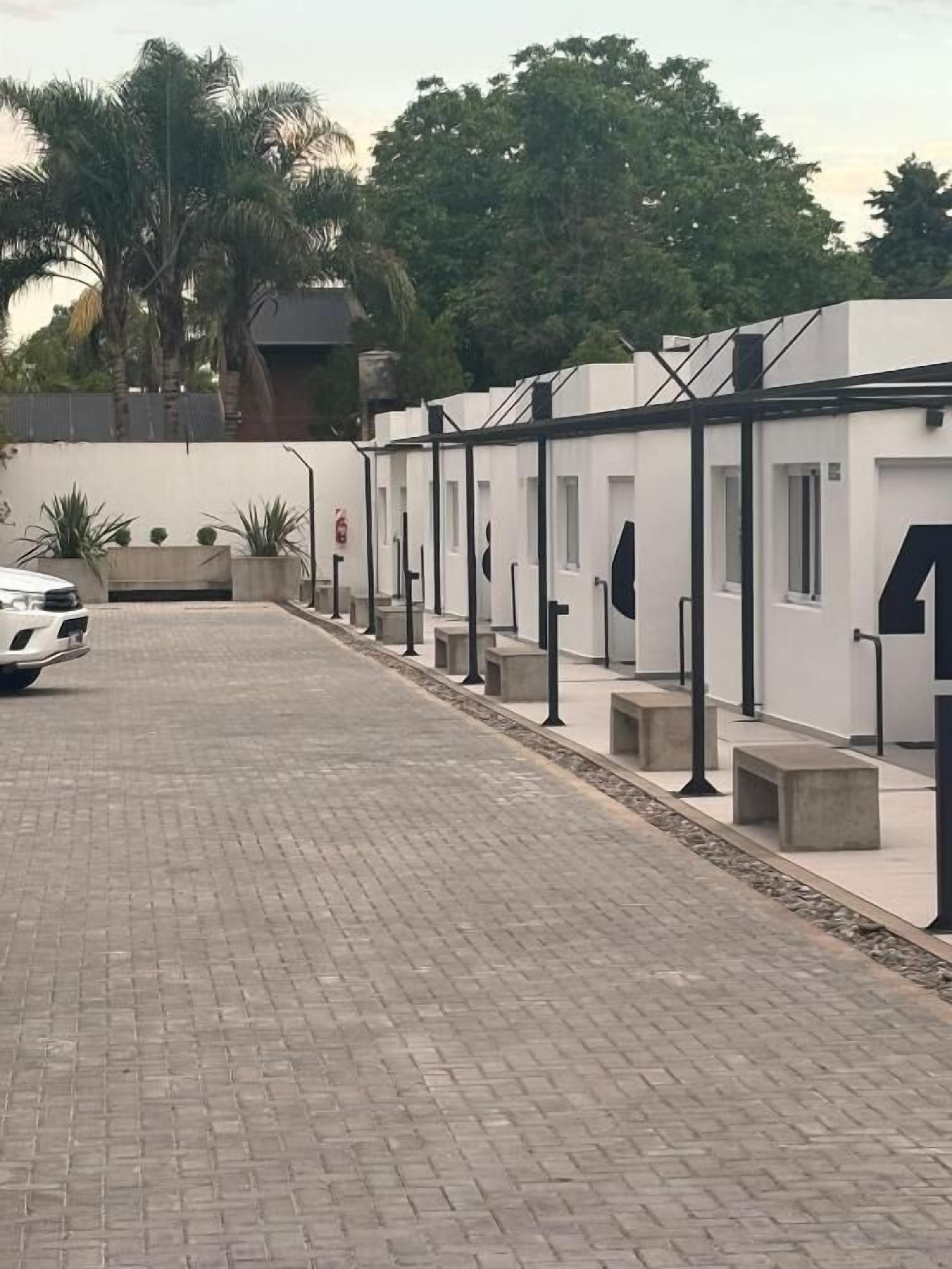 A paved pathway leading to a row of white buildings with black railings, a white car parked on the left, and a palm tree in the background.