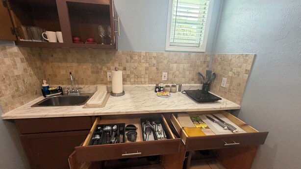 A kitchen with a marble countertop, a sink, a window with blinds, and a drawer with utensils. - The Blue Palace (Tampa)