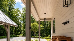 A covered wooden deck with a pergola, a stack of firewood, and a garden with white flowers. - Rabbit Ridge New Designer Cottage Screened Porch + Wood Fireplace | EV Charger (Cashiers)