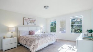 A bedroom with a bed, two bedside tables, a nightstand, a wall-mounted lamp, a ceiling light, and a window with a view of greenery. - Bella Vida 4BR Home • Private Pool & Lake View (Kissimmee)