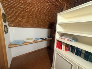 A room with a brick ceiling, a white wall, a wooden shelf with folded towels, and a white cabinet with books. - TERRAZZA PALAZZO VIALE CERVO (Cervo)