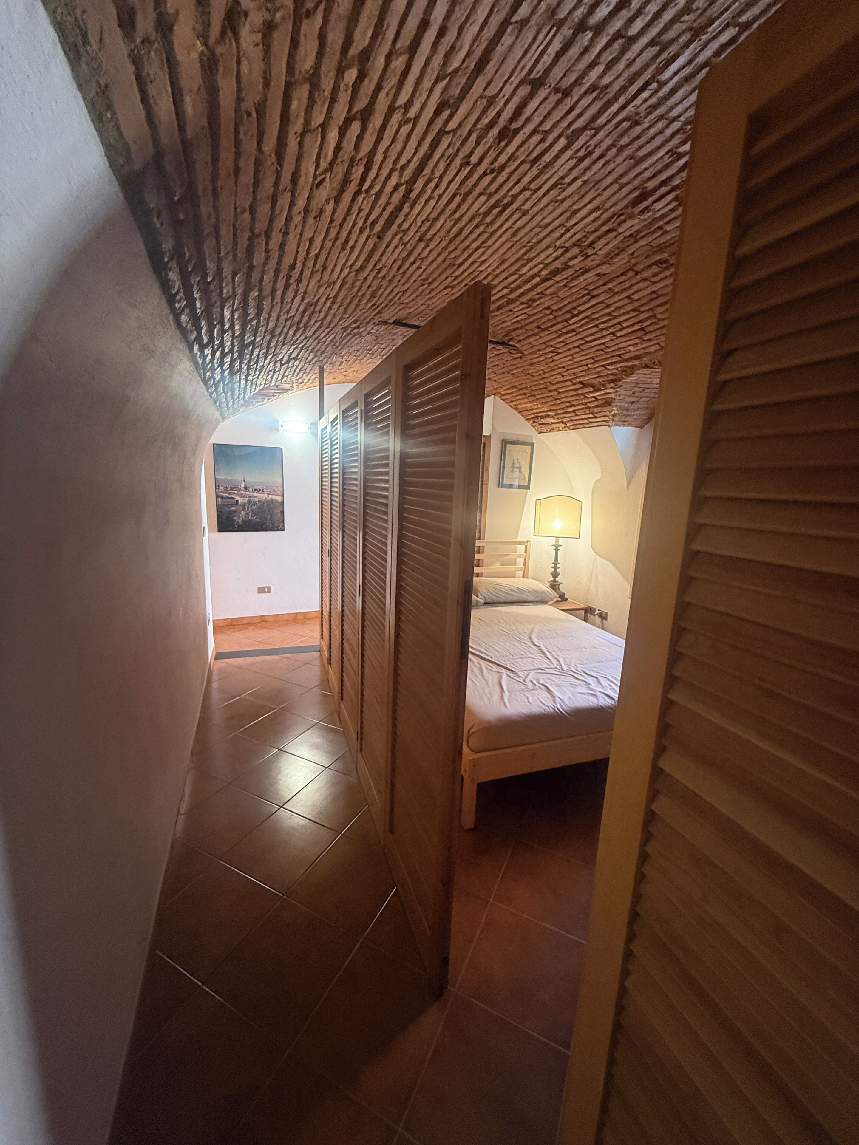 A narrow hallway with a sloped ceiling leading to a bedroom with a bed and a nightstand.