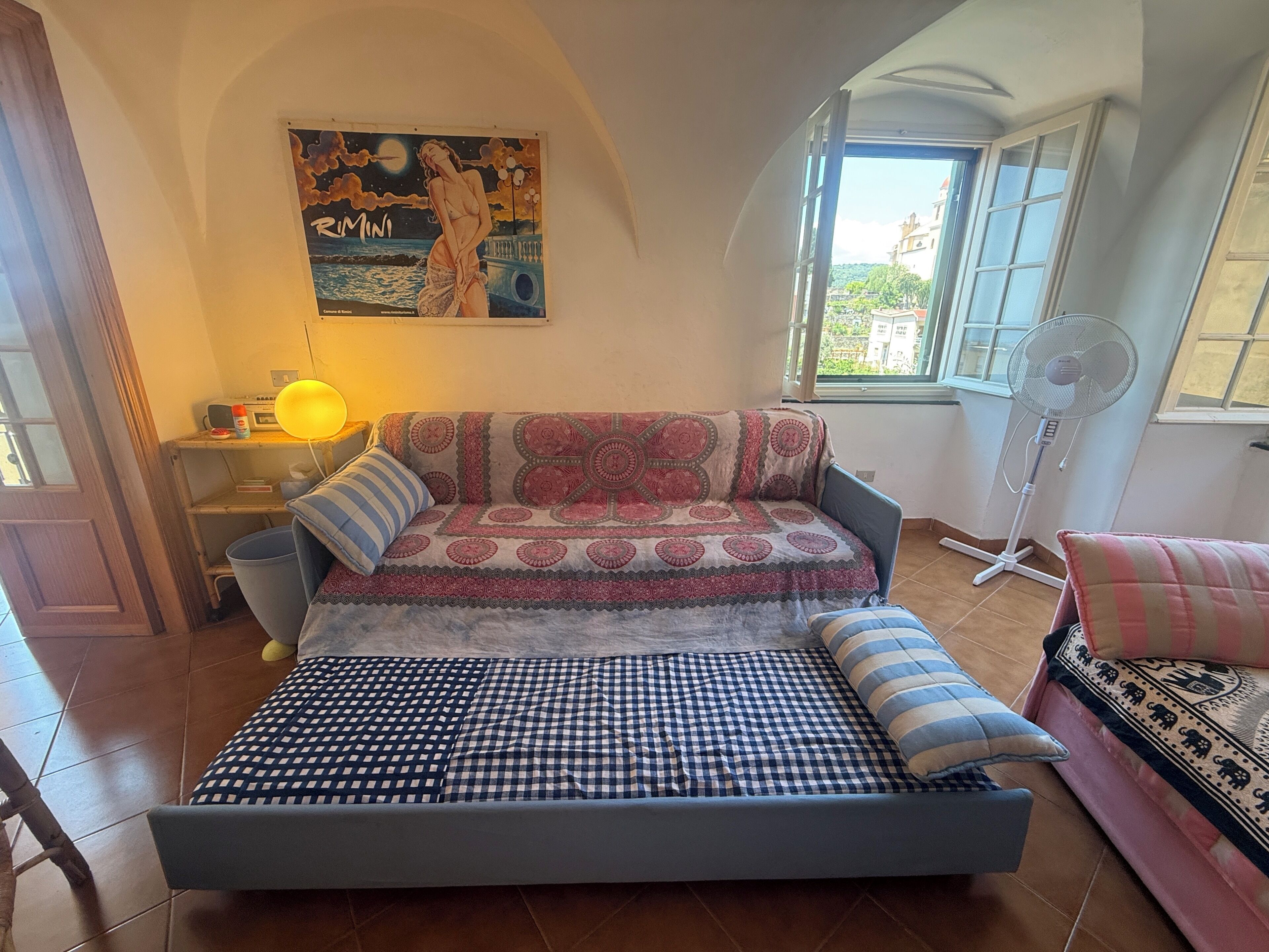 Een knus interieur met een slaapbank, een bed, een ventilator, een lamp en een schilderij aan de muur.