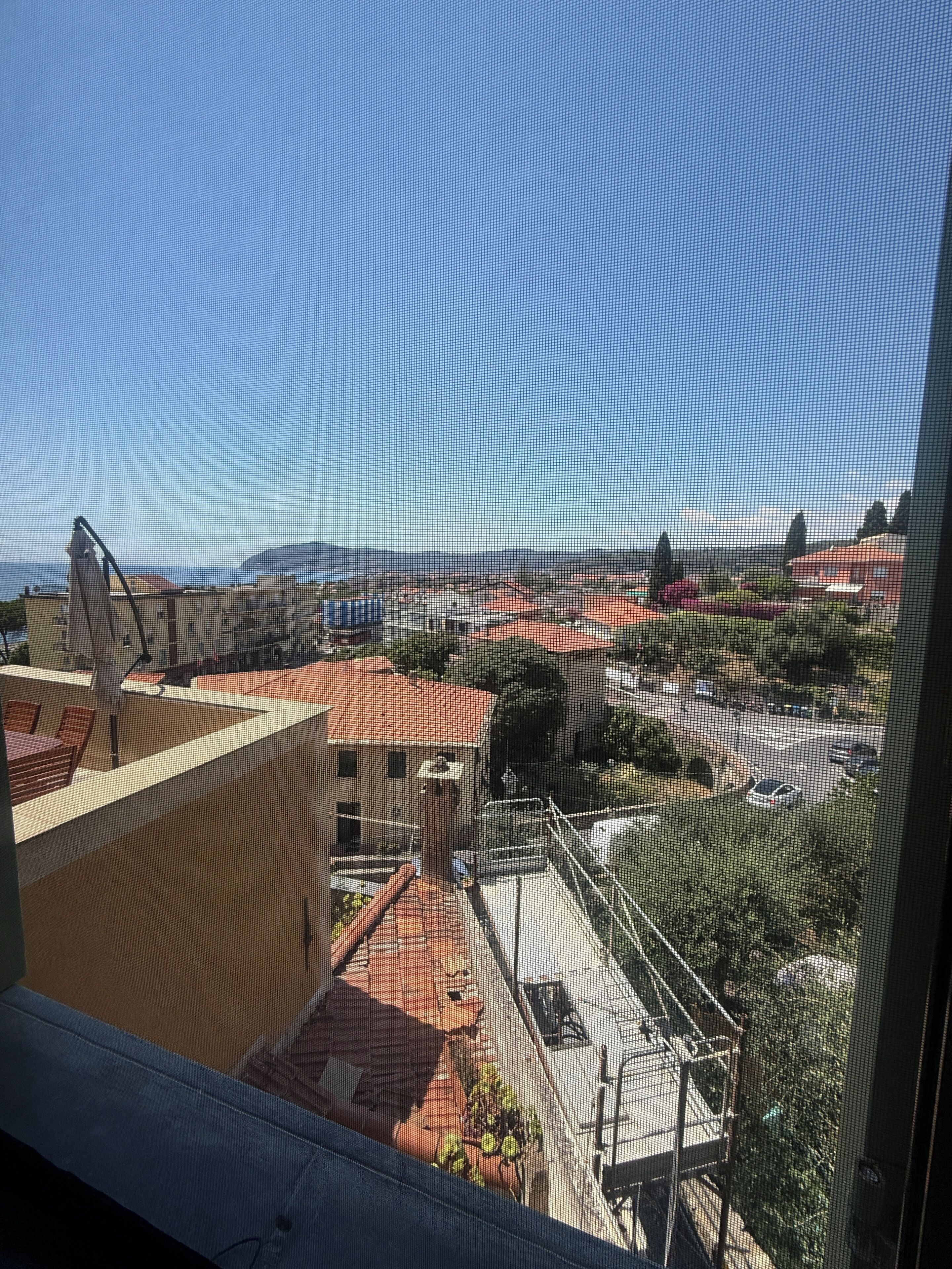 A view through a window with a mesh screen, featuring a building with a red roof, a parking area, and a clear sky.