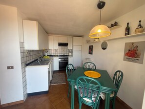 Cocina compacta con armarios blancos, estufa y pileta. Se observa una mesa de comedor con sillas y un tazón amarillo encima.