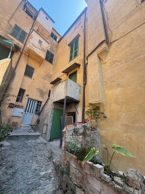 A narrow alleyway with buildings on either side, featuring a small outdoor seating area with a table and chairs, a brick wall with plants, and a green door. - TERRAZZA PALAZZO VIALE CERVO (Cervo)
