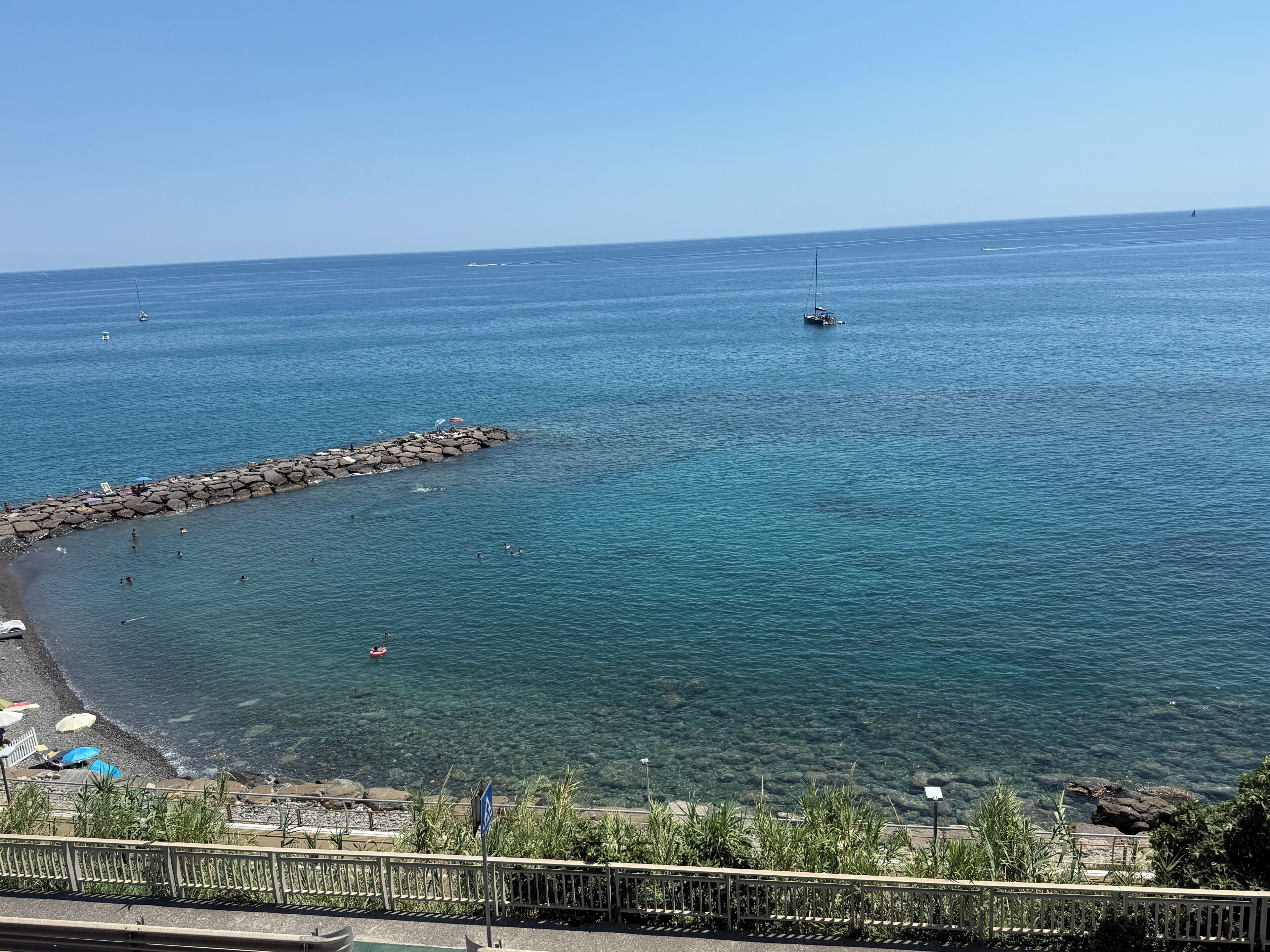Une vue sur la côte, avec un brise-lames rocheux, une eau d’un bleu limpide et quelques bateaux.