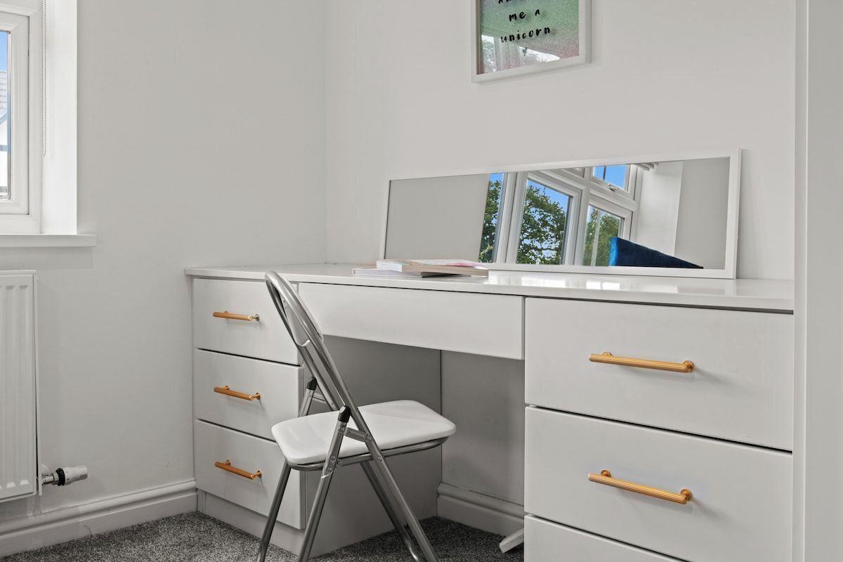 Ein weißer Schreibtisch mit Schubladen und ein Stuhl, ein Fenster und ein gerahmtes Bild an der Wand.