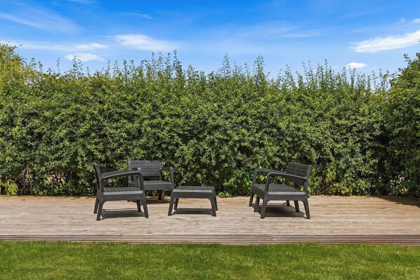 A patio with three grey chairs and a table, surrounded by a hedge.