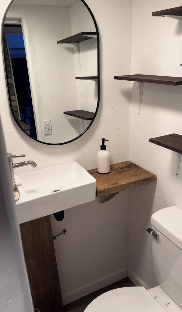 A bathroom with a white sink, a mirror, a toilet, and wooden shelves.