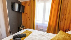 A hotel room with a bed, a flat-screen TV mounted on the wall, a small table with a statue, and a window with curtains.