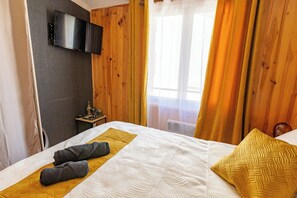 Habitación de hotel con cama, televisor de pantalla plana montado en la pared, una mesita con una estatua y una ventana con cortinas.