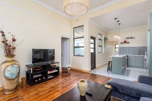 Una sala de estar con televisor, una mesa de centro y un sofá. Hay una zona de cocina con un lavamanos y armarios.