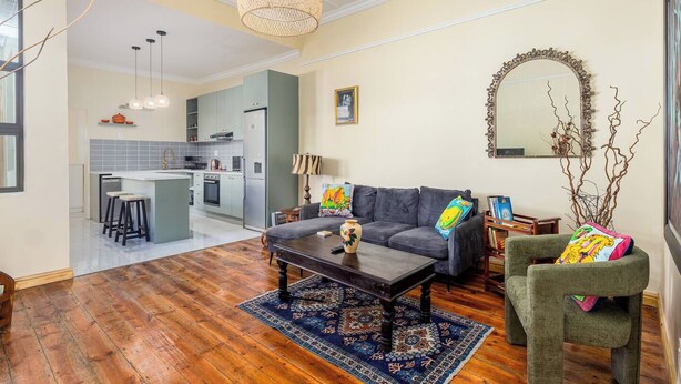 Una sala de estar moderna con sofá, mesa de centro y alfombra. Se observa una cocina al fondo con comedor.