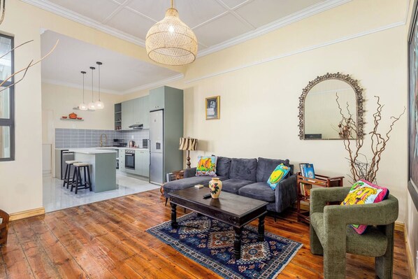Una sala de estar moderna con sofá, mesa de centro y alfombra. Se observa una cocina al fondo con comedor.