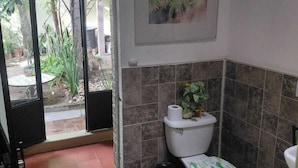 A bathroom with a toilet, a sink, and a shower area. There is a door leading to an outdoor area with plants. - Casa Aldama 83. (San Miguel de Allende)
