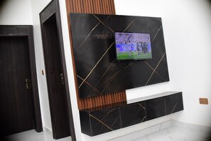 A room with a dark wooden door, a black wall with a TV mounted above a black marble shelf, and a white wall with a wooden panel.