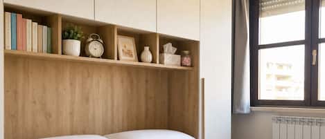 Un lit bien fait, avec du linge de lit et des serviettes blanches, une tête de lit en bois et une étagère contenant des livres, une horloge et des objets décoratifs.