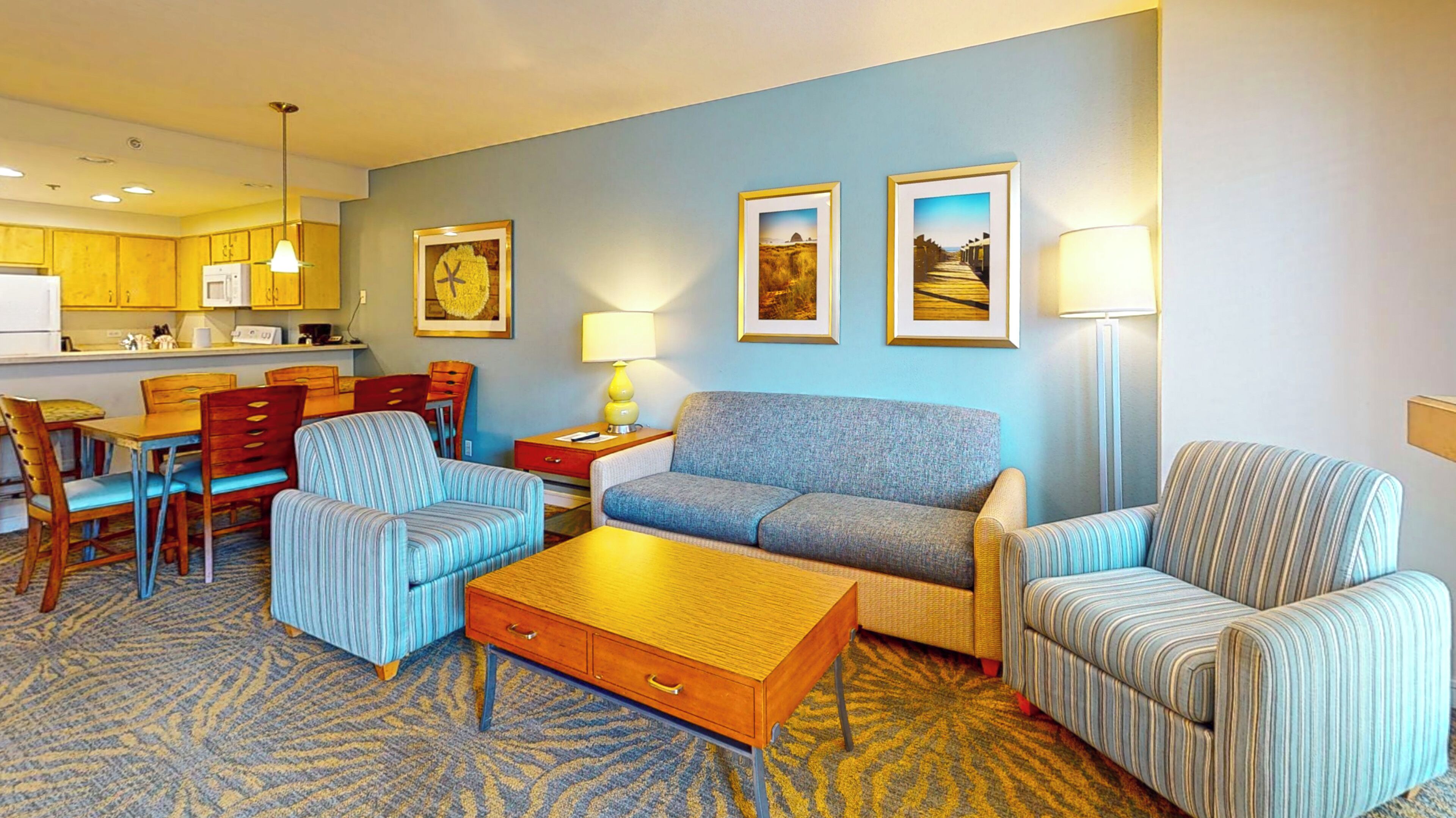 A hotel room with a dining area, a sofa set, a lamp, and framed pictures on the wall.