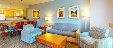 A hotel room with a dining area, a sofa set, a lamp, and framed pictures on the wall.