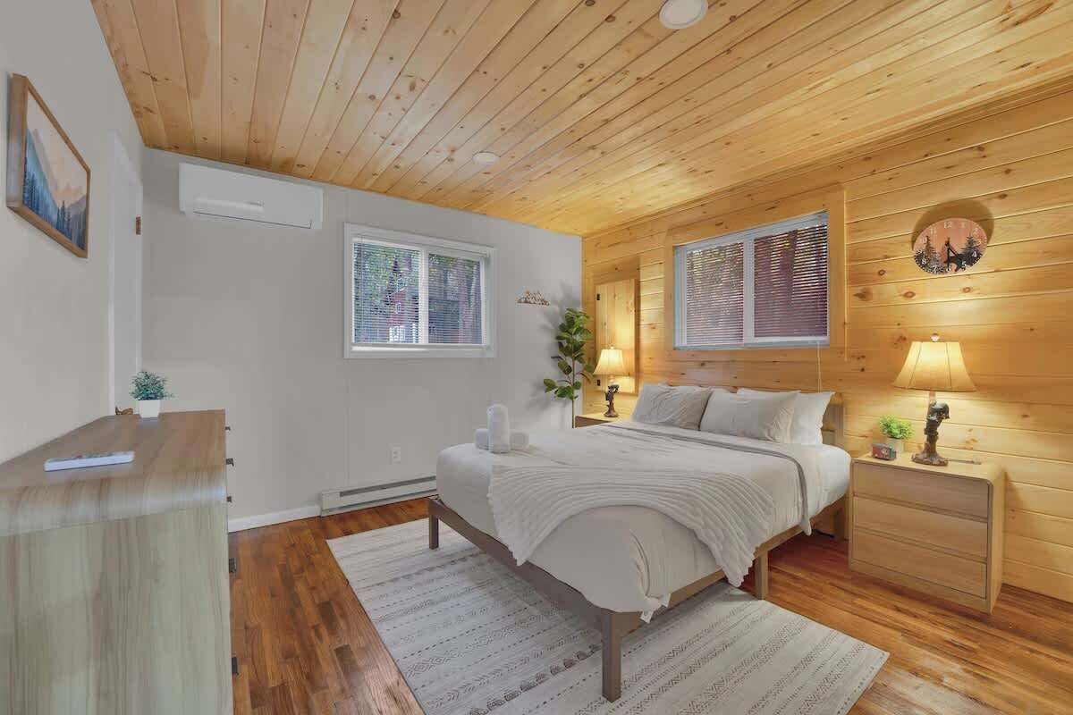 A bedroom with a wooden ceiling, a bed with white bedding, two bedside tables with lamps, and a small plant on one table.