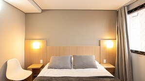 Hotellihuone, jossa on sänky, kaksi yöpöytälamppua, tuoli ja ikkuna, jossa on verhot.
