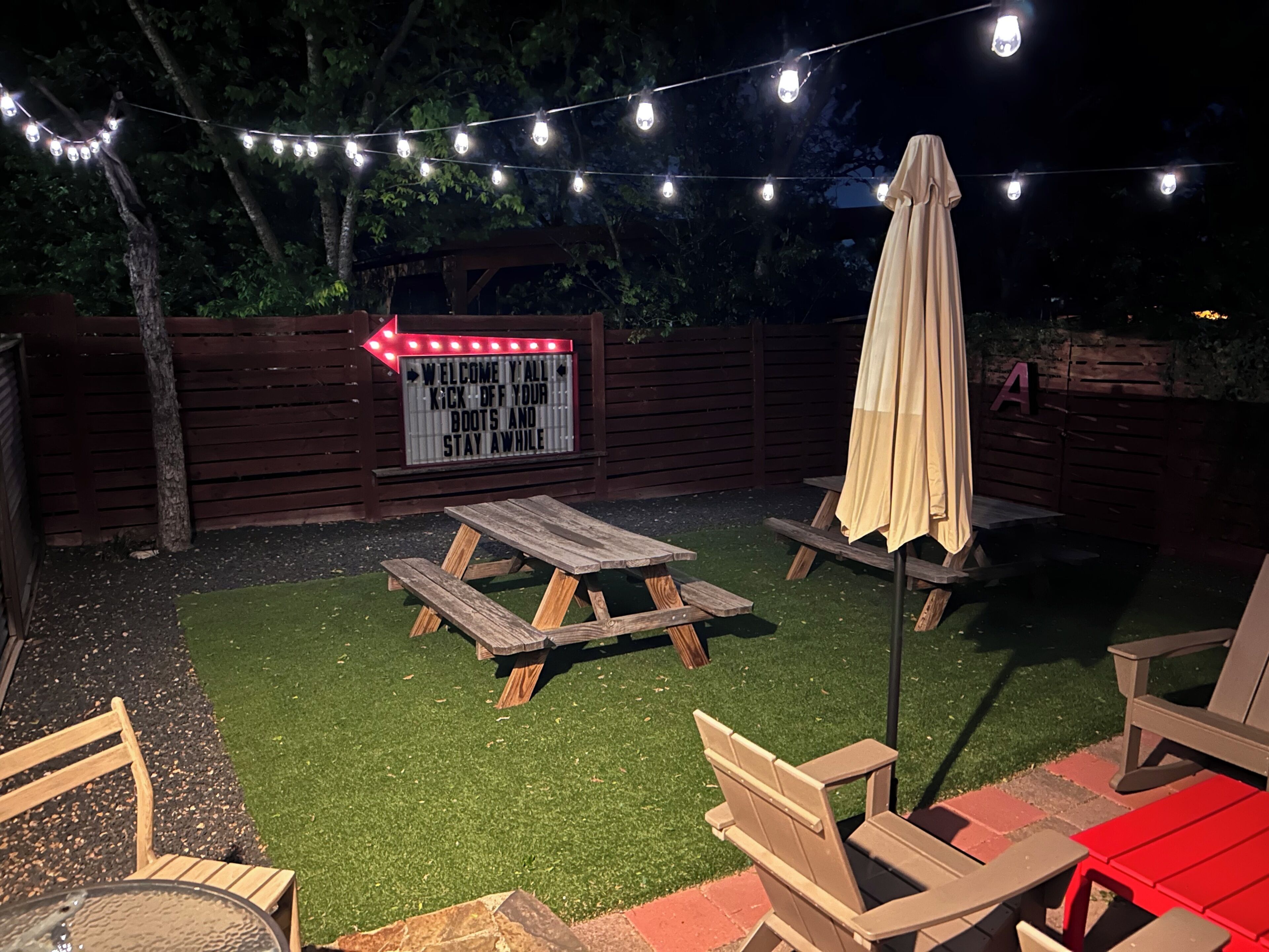 A backyard with artificial grass, wooden picnic tables, and string lights.