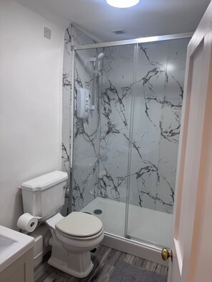 A bathroom with a toilet, a shower enclosure, and a marble wall. - Stony Stratford Cozy Apartment (Milton Keynes)