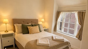 A neatly made bed with pillows and a folded towel, a bedside table with a lamp, and a window with curtains. - Stony Stratford Cozy Apartment (Milton Keynes)