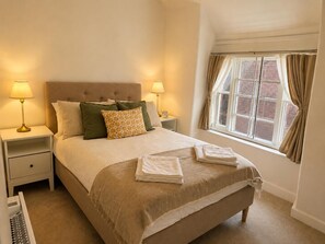 A neatly made bed with pillows and a folded towel, a bedside table with a lamp, and a window with curtains. - Stony Stratford Cozy Apartment (Milton Keynes)