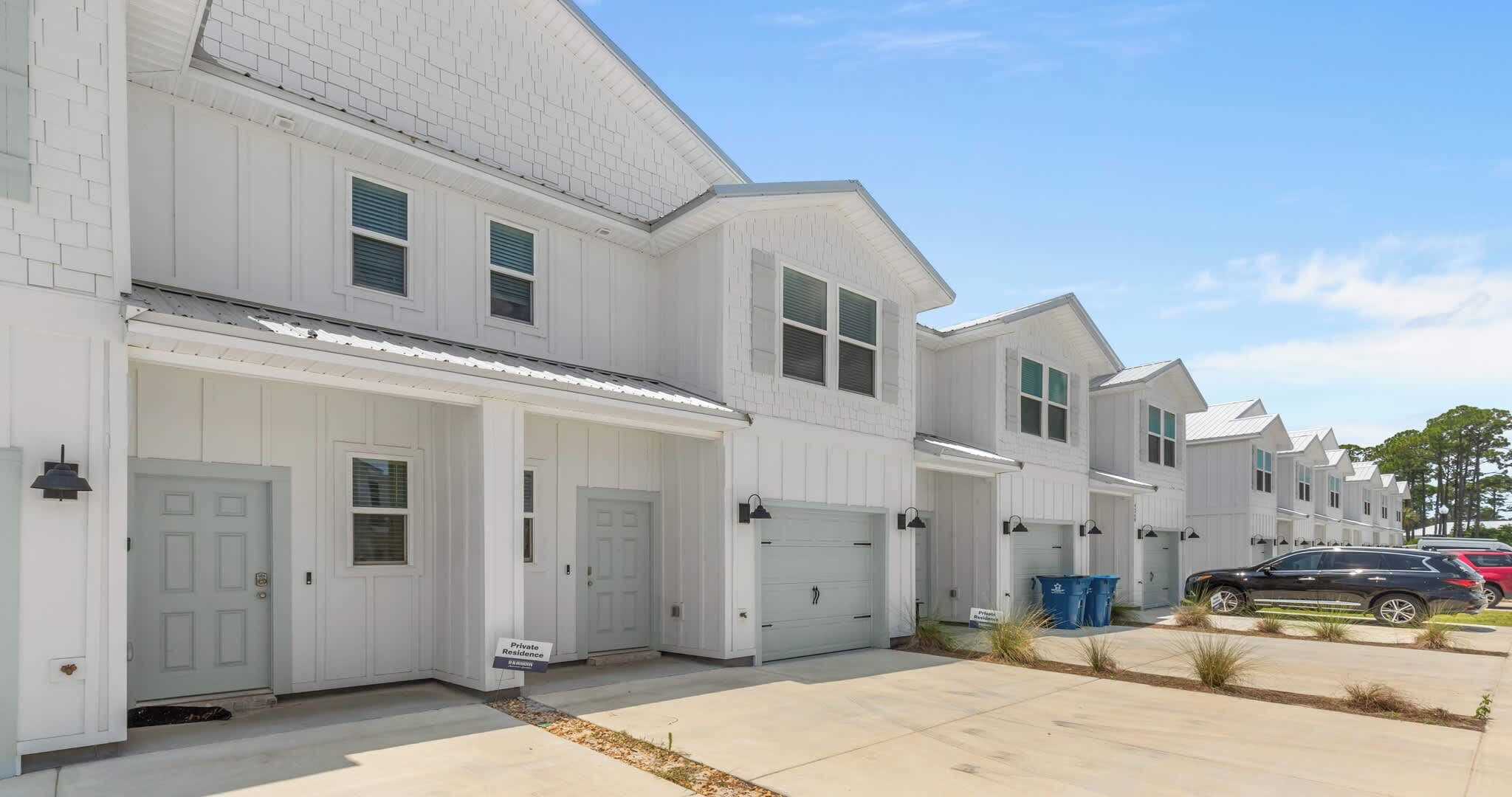 A row of white single-family homes with attached garages, a clear sky, and a parking area with cars.