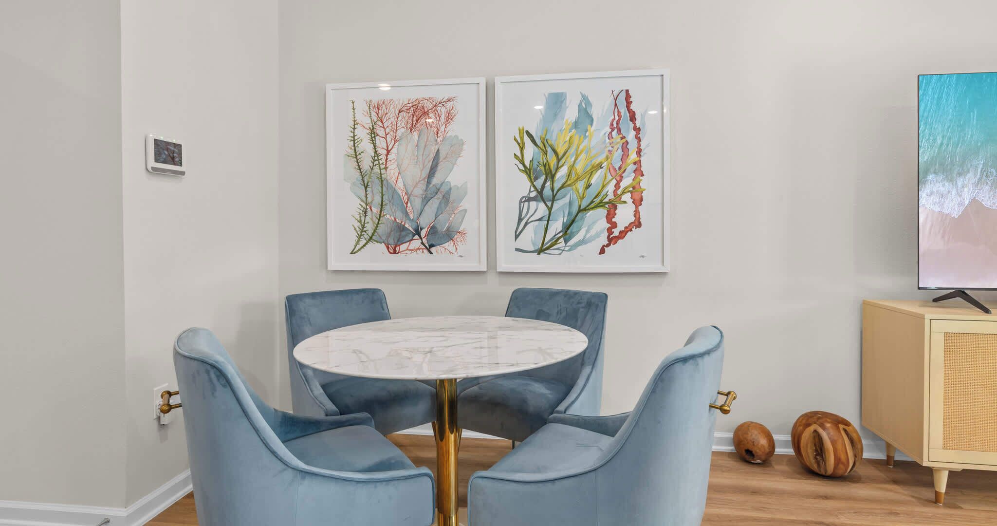 A dining area with a round table, four blue chairs, and two framed botanical prints on the wall.