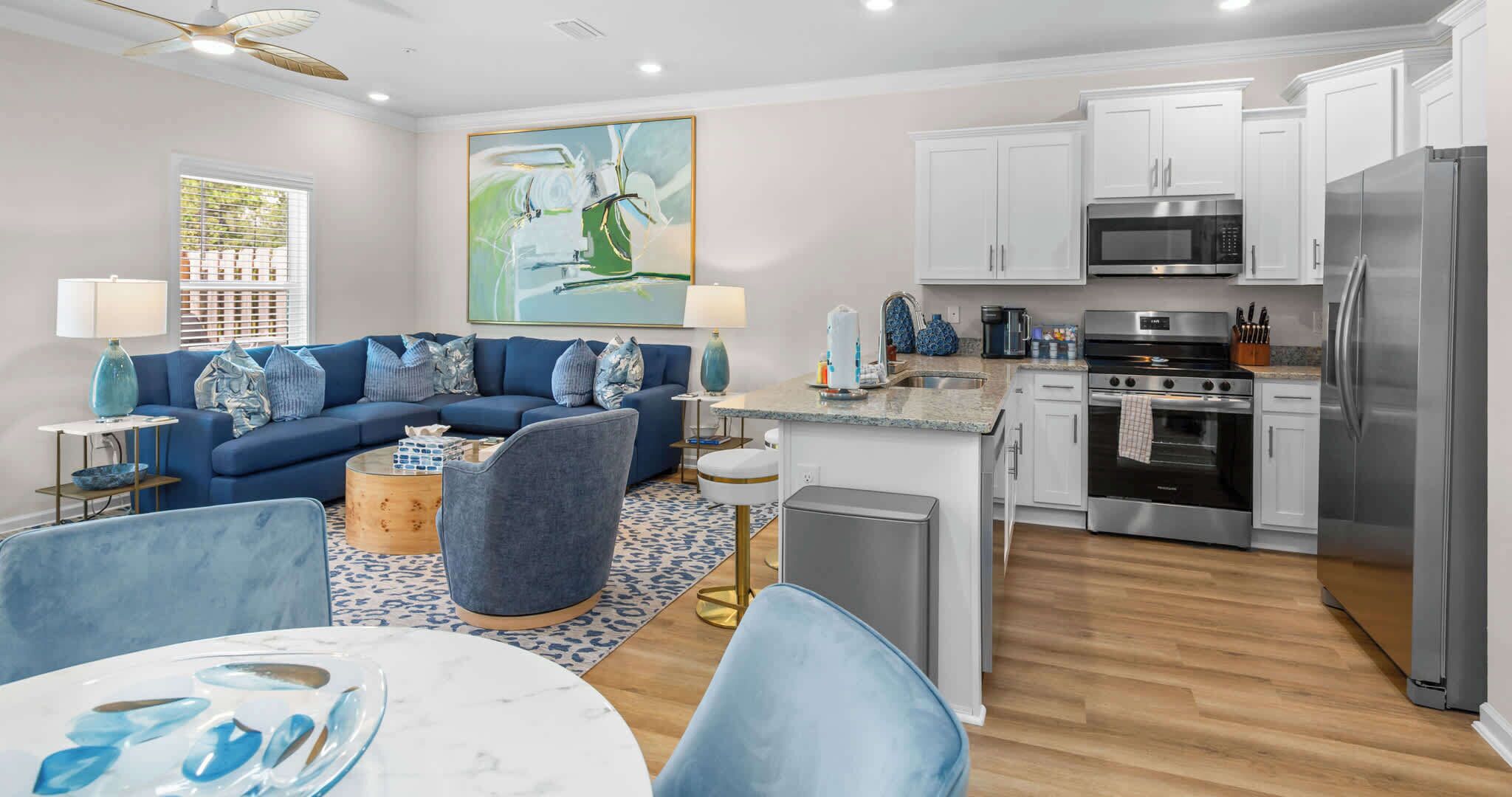 A modern living room with a blue sofa, a dining area with a round table, and a kitchen with white cabinets and stainless steel appliances.
