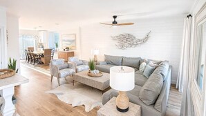 Ein geräumiges Wohnzimmer mit einem grauen Sofa, einem Couchtisch und einer Lampe. Im Hintergrund ist eine Essecke mit Tisch und Stühlen zu sehen.