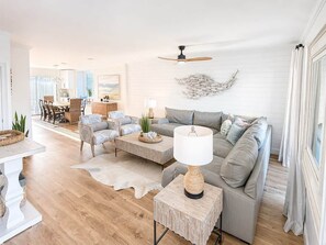 A spacious living room with a grey sofa, a coffee table, and a lamp. There is a dining area in the background with a table and chairs. - Gulf Winds East #12 - Pearl of the Sea (Miramar Beach)