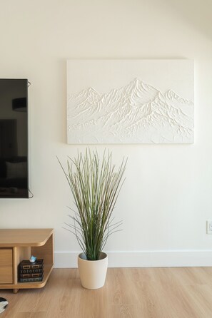 A room with a flat-screen TV, a wall-mounted artwork depicting a mountain landscape, a potted plant, and a wooden cabinet with books. - Luxury Escape, Backyard Pool & Hillside Views (Los Angeles)