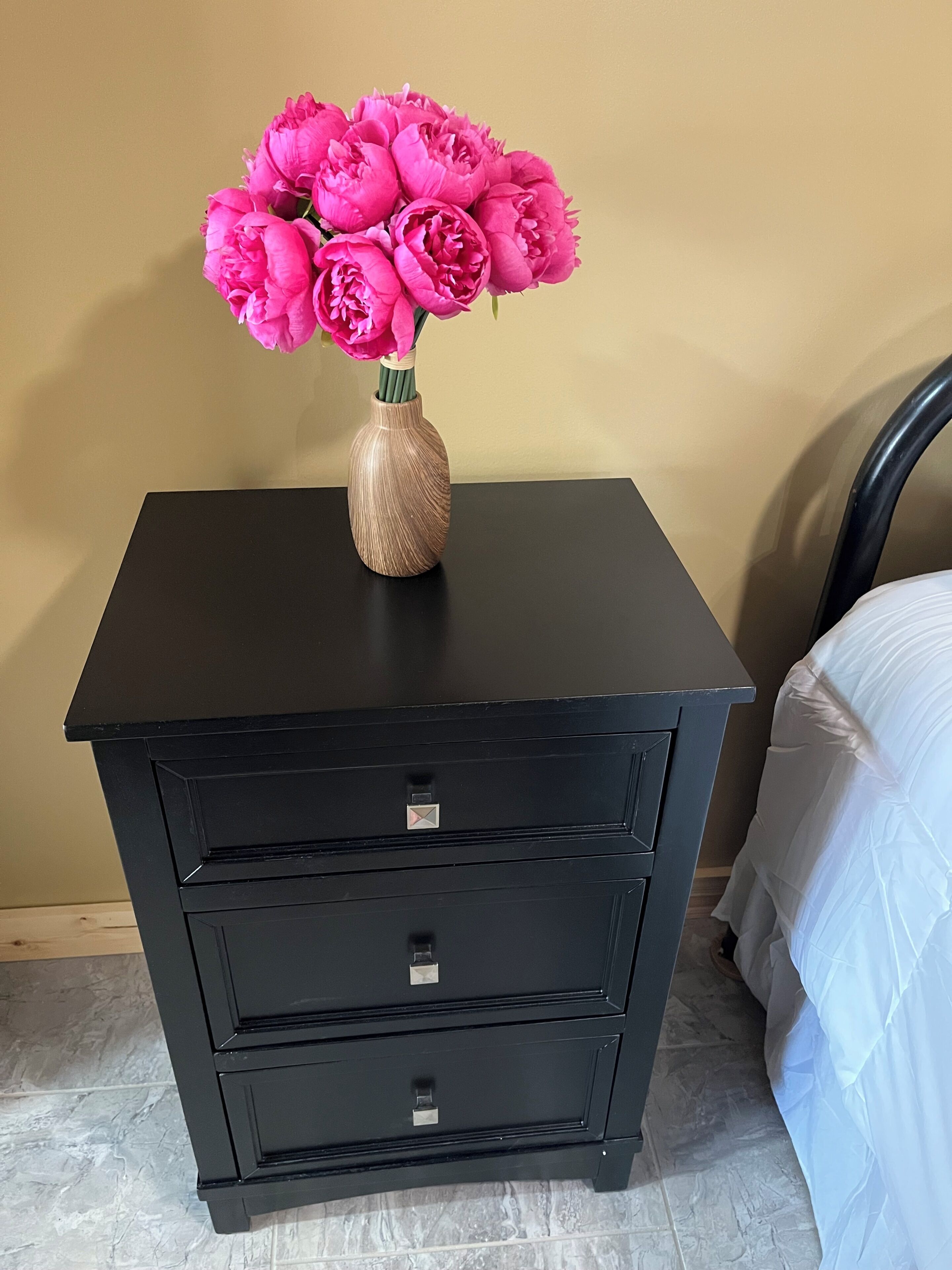 A black nightstand with a vase of pink flowers, a drawer, and a bed with a white bedspread.