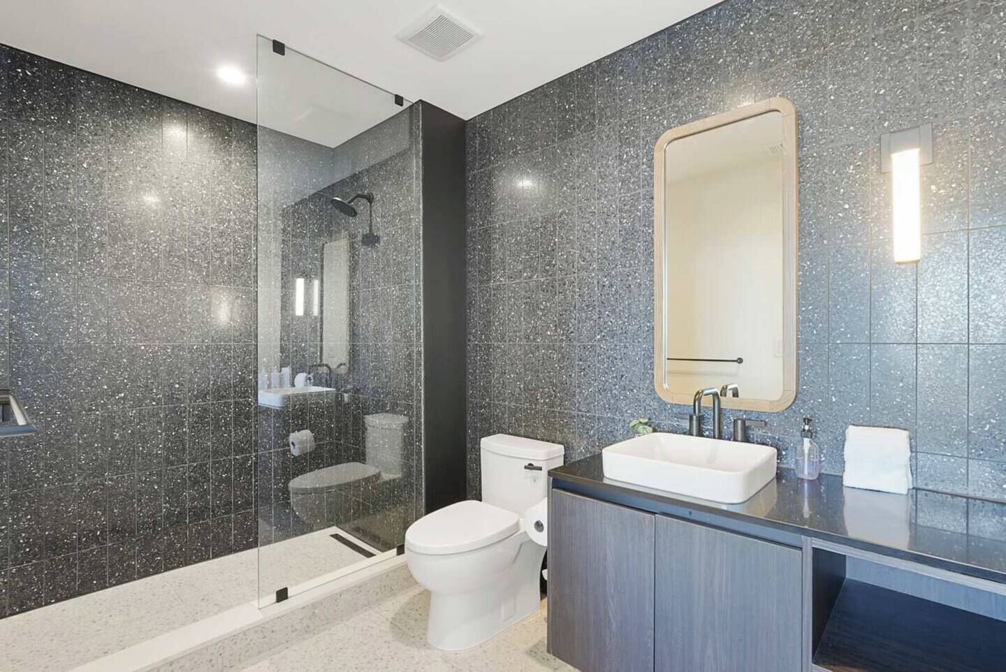 Un baño moderno con cabina de ducha de vidrio, inodoro, lavamanos con espejo y pared revestida de baldosas oscuras.