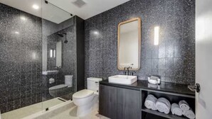 A modern bathroom with a glass shower enclosure, a white toilet, a sink with a mirror, and a towel rack with towels. - Luxury High-Rise 2BR (Austin)