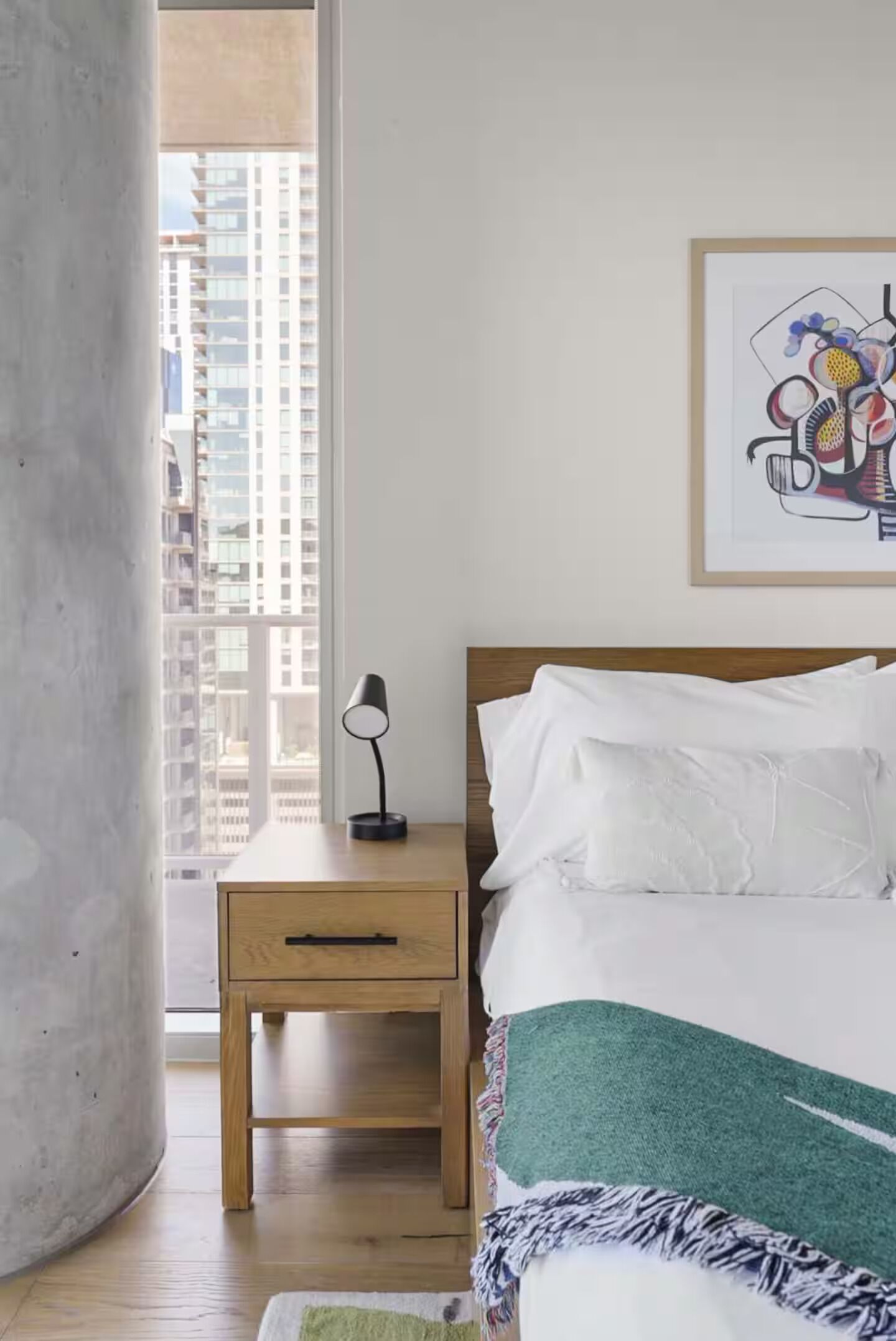 Un dormitorio con cama, mesita de noche, lámpara, una obra de arte enmarcada y vistas a edificios a través de una ventana.