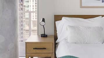 Un dormitorio con cama, mesita de noche, lámpara, una obra de arte enmarcada y vistas a edificios a través de una ventana.
