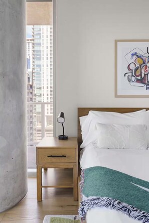 A bedroom with a bed, a nightstand, a lamp, a framed artwork, and a view of buildings through a window. - Luxury High-Rise 2BR (Austin)