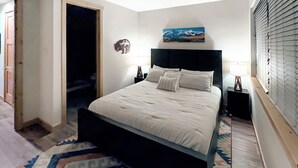 Quarto com cama, mesa de cabeceira, abajur, um quadro na parede e uma porta para outro cômodo.