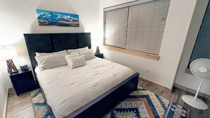 En seng med hvide sengetøj, en mørk sengegavl, et vindue med persienner, et billede over sengen, et natbord med en lampe og en gulvventilator.